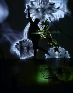 This image appears to be an artistic photograph featuring a person silhouetted against a backdrop of illuminated, abstract floral visuals. The setting likely represents a performance or art installation titled "Learning to Move, Learning to Play, Learning to Animate" (2024), with the photograph credited to the Cali2 AV team. The scene blends organic elements (plants in the foreground and possibly attached to the performer) with digital projections, creating a fusion of natural and technological aesthetics. The moody lighting and dynamic pose suggest themes of interaction, growth, or transformation.