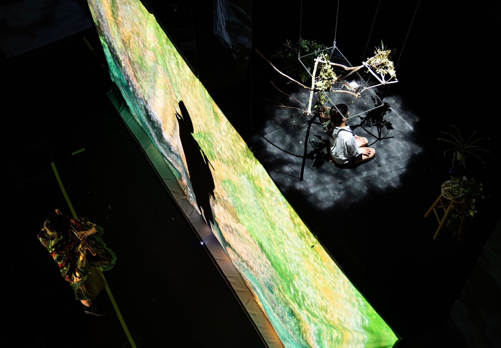 A photograph from the multimedia performance "Learning to Move, Learning to Play, Learning to Animate" (2024), captured by the Cali2 AV team. The scene is viewed from above, showing one performer standing to the left in colorful attire and another performer seated on the floor to the right, surrounded by suspended branches and plants. A large, brightly illuminated screen divides the space, casting dramatic shadows and highlighting the contrast between light and dark areas on stage.
